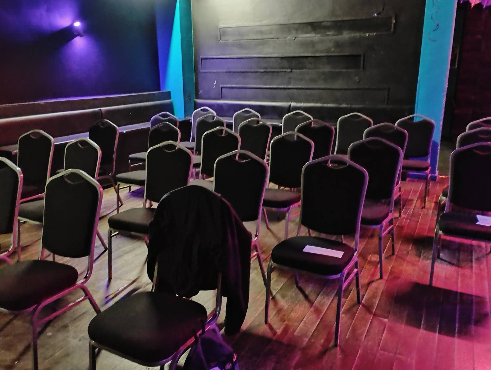 a smallish room with a wooden floor and black panelled walls filled with black chairs, some which have handouts on them.